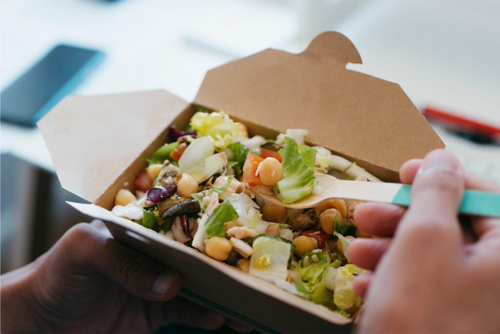 Sharebite-Feature-1253x836 A person holds a cardboard takeout container filled with salad, using a fork to eat. The salad contains lettuce, chickpeas, beans, and other fresh vegetables.