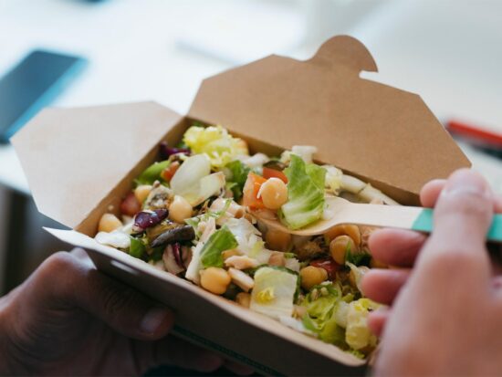 A person holds a cardboard takeout container filled with salad, using a fork to eat. The salad contains lettuce, chickpeas, beans, and other fresh vegetables.