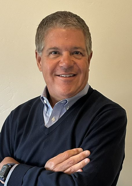 A middle-aged man with short gray hair, wearing a navy blue sweater over a light blue collared shirt, smiles with his arms crossed against a plain light-colored wall.