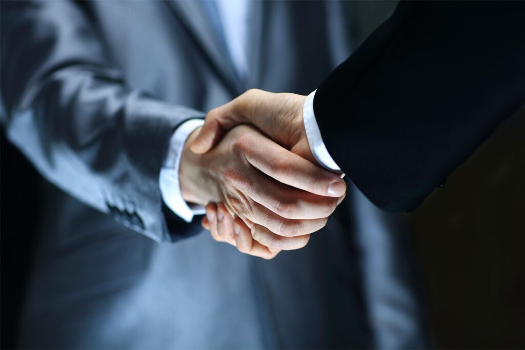 Two people wearing business suits are shaking hands, symbolizing agreement or partnership, with the focus on their hands and blurred upper bodies in the background.