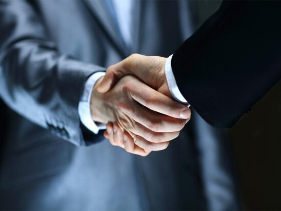 Two people wearing business suits are shaking hands, symbolizing agreement or partnership, with the focus on their hands and blurred upper bodies in the background.