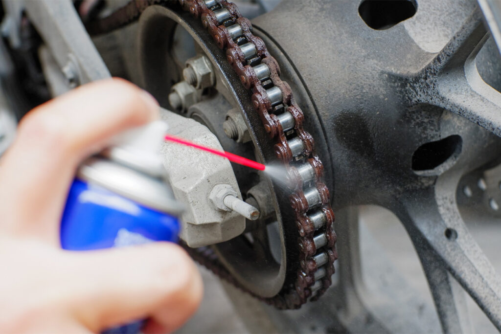 WD-40-Spray A person sprays lubricant onto a motorcycle chain, focusing on the sprocket and chain area to ensure smooth operation and prevent rust.