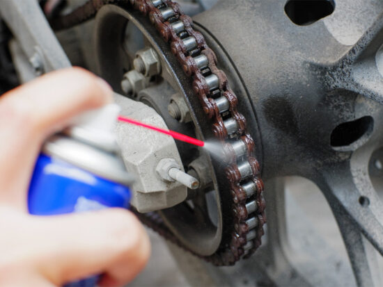 A person sprays lubricant onto a motorcycle chain, focusing on the sprocket and chain area to ensure smooth operation and prevent rust.