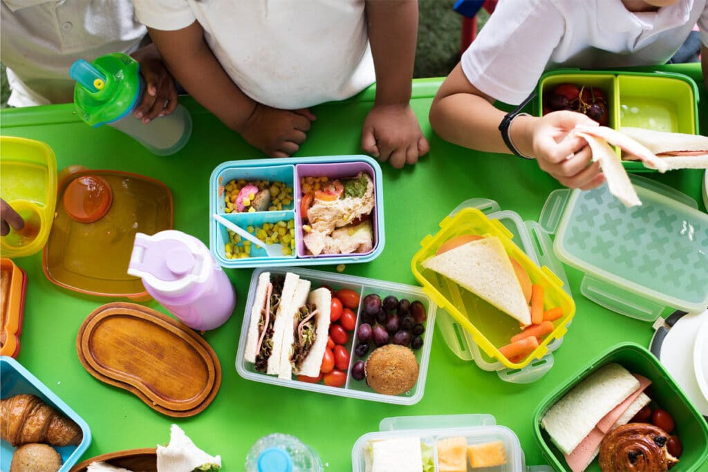 GBC-Revolution-Foods-Thumbnail Overhead view of several children eating lunch from colorful plastic containers filled with sandwiches, fruit, vegetables, and snacks at a bright green table.
